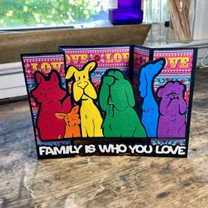 Peut inclure: Une carte à panneaux multiples colorée avec des silhouettes de chiens arc-en-ciel. La carte a un fond rose avec le mot "LOVE" répété. La phrase "FAMILY IS WHO YOU LOVE" est affichée en bas.