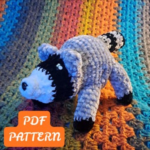 Puede incluir: Un juguete de peluche de mapache tejido a ganchillo con hilo gris suave, con una máscara negra en los ojos, un hocico blanco, una nariz negra y una cola a rayas. Las cuatro patas tienen puntas negras. Un gráfico naranja muestra el texto PDF PATTERN.