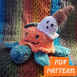 May include: A plush crochet turtle with a teal body and a bright orange shell. A small white ghost with a brown pointed hat sits on top of the shell. Both the turtle and the ghost have black embroidered eyes and mouths. Includes a PDF PATTERN label.