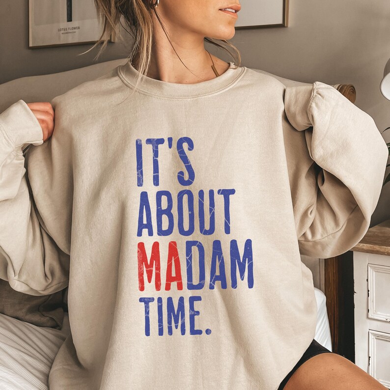 Może przedstawiać: Beżowa bluza z nadrukiem "IT'S ABOUT MADAM TIME." w niebieskich i czerwonych literach.
