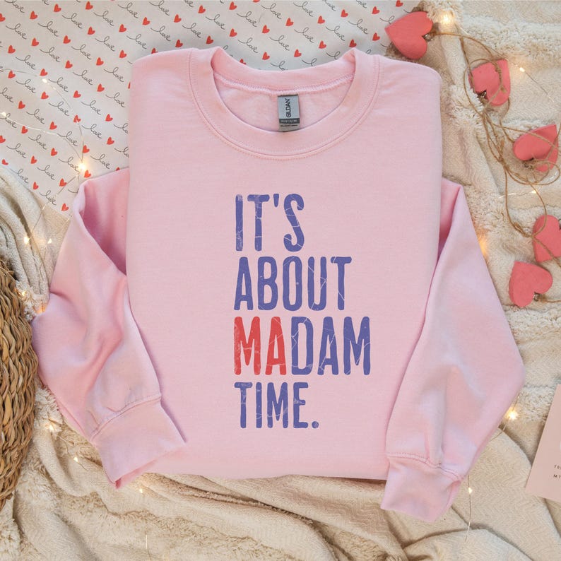 Może przedstawiać: R&oacute;żowa bluza z nadrukiem "IT'S ABOUT MADAM TIME." w kolorze niebieskim i czerwonym.