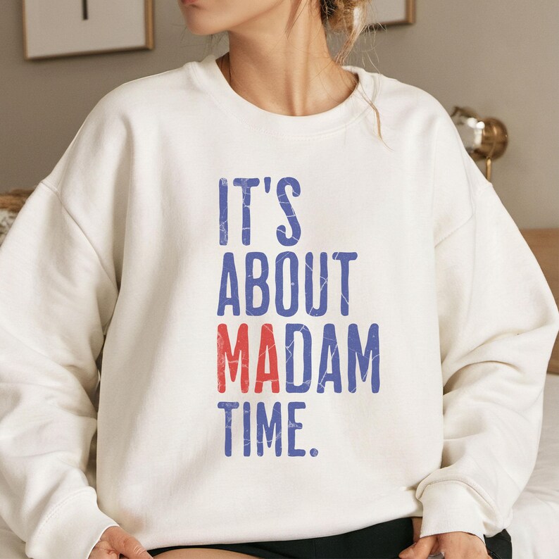 Może przedstawiać: Biała bluza z napisem "It's About Madam Time." w niebieskich i czerwonych literach.