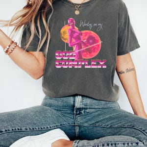 May include: Dark gray t-shirt with a graphic design featuring a pink figure lifting weights, a pink and orange planet, and the text "Working on my God Complex." The shirt is a relaxed fit.