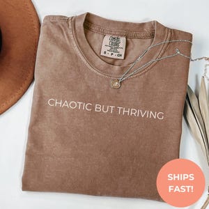 May include: A brown t-shirt with the text "CHAOTIC BUT THRIVING" printed in white on the front.