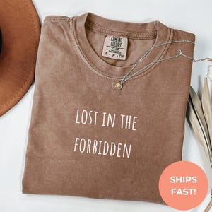 May include: A brown t-shirt with the text "LOST IN THE FORBIDDEN" in white. The shirt has a crew neck and short sleeves. A silver necklace with a pendant is draped across the shirt. The shirt is folded on a white surface.