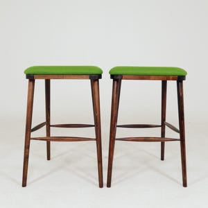 May include: Two wooden stools with green upholstered seats. The stools have dark brown, slender legs and a horizontal support bar. The seats are rectangular and the green fabric contrasts with the wood.