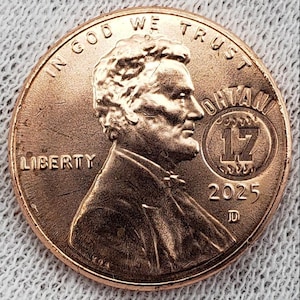Puede incluir: Una moneda de un centavo de color cobre con el perfil de un hombre, la palabra "LIBERTY" y el año "2025". La moneda también presenta el texto "IN GOD WE TRUST" y la palabra "OHIO" con el número "17".