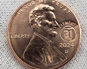May include: A copper-colored US penny coin with the words "In God We Trust" and "Liberty" inscribed on it. The coin features a profile of Abraham Lincoln and a commemorative design for "Clemente 21" with the year 2024 and the letter "D" inscribed on the coin.
