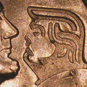 May include: Close-up of a copper-colored coin featuring two profile portraits. One profile is of a man with a prominent nose, and the other is a woman with stylized hair. The coin's surface has a textured appearance.