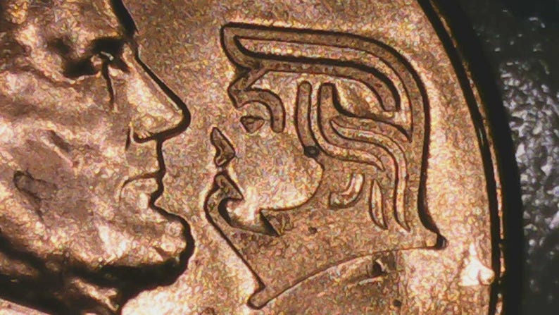 May include: Close-up of a copper-colored coin featuring two profile portraits. One profile is of a man with a prominent nose, and the other is a woman with stylized hair. The coin's surface has a textured appearance.
