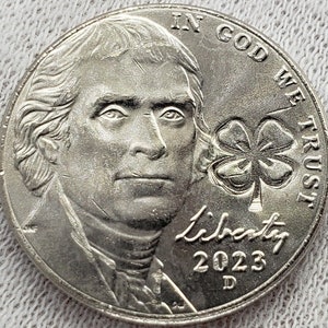 May include: A silver US nickel coin with the profile of Thomas Jefferson on the obverse and a four-leaf clover with the inscription "In God We Trust" on the reverse. The coin is dated 2023 and has the mint mark "D".