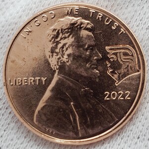May include: A copper-colored 2022 penny with the profile of Abraham Lincoln on the obverse. The coin features the inscriptions "IN GOD WE TRUST" and "LIBERTY". A second profile is etched on the coin.