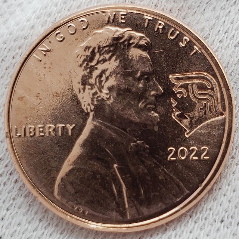 May include: A copper-colored 2022 penny with the profile of Abraham Lincoln on the obverse. The coin features the inscriptions "IN GOD WE TRUST" and "LIBERTY". A second profile is etched on the coin.