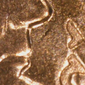 May include: Close-up of a copper-colored coin, showing intricate details of a raised design. The surface has a textured appearance with visible lines and shapes, suggesting a historical or collectible item.