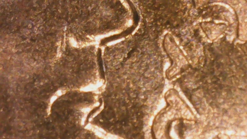 May include: Close-up of a copper-colored coin, showing intricate details of a raised design. The surface has a textured appearance with visible lines and shapes, suggesting a historical or collectible item.