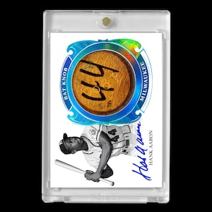 May include: A baseball card featuring Hank Aaron in a Milwaukee Brewers uniform, holding a bat. The card includes a bat knob with the number 44 and the text "Bat Knob Milwaukee". The card is encased in a clear protective holder.