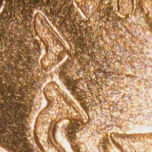 May include: Close-up of a gold-colored coin, showing detailed relief patterns. The surface has a textured appearance with intricate designs and shapes. The coin's design includes a profile and other abstract elements.