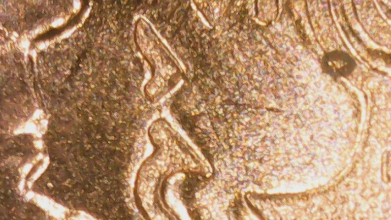 May include: Close-up of a gold-colored coin, showing detailed relief patterns. The surface has a textured appearance with intricate designs and shapes. The coin's design includes a profile and other abstract elements.