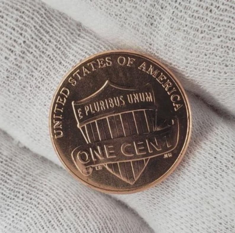 May include: A copper-colored United States one-cent coin. The coin features the words "UNITED STATES OF AMERICA" and "ONE CENT". The coin also has the words "E PLURIBUS UNUM" and a shield design.