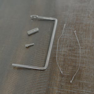 May include: A selection of silver-coloured hardware components, including a bent metal tool, two small cylindrical pieces, and two thin, white strings with small, round ends. The items are arranged on a wooden surface.