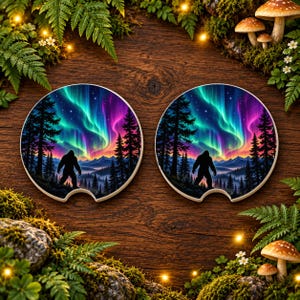 May include: Two round car coasters featuring a vibrant illustration of a silhouette of a Bigfoot creature standing in a forest of pine trees against a backdrop of colorful aurora borealis lights, starry night sky, and distant mountain peaks.