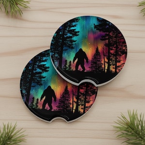 Puede incluir: Dos posavasos redondos para coche con una silueta de Bigfoot en una escena de bosque bajo una colorida aurora boreal. Los posavasos tienen un borde negro y una base blanca, perfectos para absorber derrames. El diseño incluye árboles y un cielo vibrante.