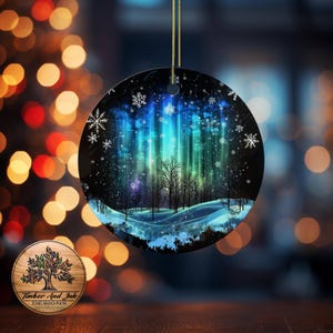 Puede incluir: Adorno navideño redondo con una escena invernal que presenta un bosque y la aurora boreal. El adorno tiene un fondo negro con colores azules, verdes y blancos. Los copos de nieve están dispersos por la imagen. Se adjunta una cuerda dorada para colgar.