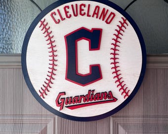 Könnte beinhalten: Rundes Holzschild mit Baseball-Design und dem Text "CLEVELAND Guardians". Das Schild hat einen weißen Hintergrund mit roten Nähten, einen marineblauen Rand und ein großes "C"-Logo in Marineblau und Rot.