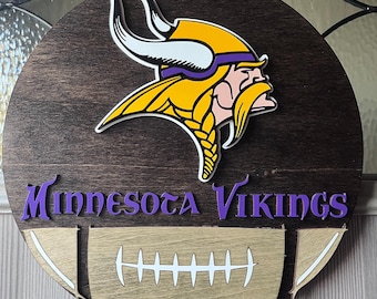 Könnte beinhalten: Ein rundes Holzschild mit dem Logo der Minnesota Vikings. Das Logo zeigt einen lila und gelben Wikingerhelm mit Hörnern und einem Football. Der Text "Minnesota Vikings" ist in lila Buchstaben unter dem Logo geschrieben.