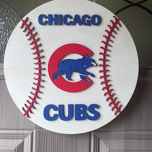 Puede incluir: Un colgador de puerta de madera blanco con forma de pelota de béisbol con costuras rojas. El logotipo de los Chicago Cubs está en el centro con una "C" roja y un oso azul. Las palabras "CHICAGO" y "CUBS" están en letras azules.