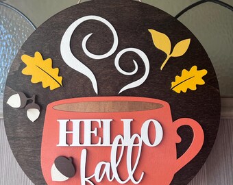 Könnte beinhalten: Ein rundes Holzschild mit einer roten Kaffeetasse, auf der der Text "HELLO fall" in Weiß steht. Aus der Tasse steigt Dampf auf und es befinden sich gelbe Blätter und Eicheln drumherum.