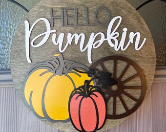 Könnte beinhalten: Ein rundes Holzschild mit dem Text "Hello Pumpkin" in weißer und grauer Schrift. Das Schild ist mit drei ausgeschnittenen Kürbissen verziert, einem gelben, einem orangen und einem braunen, sowie einem braunen Wagenrad.