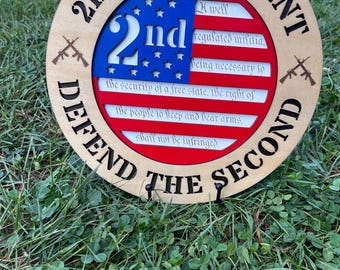 Könnte beinhalten: Runder Holzschild mit der Aufschrift "2nd AMENDMENT DEFEND THE SECOND". In der Mitte befindet sich ein blau-weißes Design mit der Zahl "2nd" und dem Text des Zweiten Zusatzartikels, mit gekreuzten Gewehren an den Seiten.