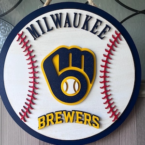 Op de afbeelding: Een rond houten bord met het Milwaukee Brewers-logo. Het bord heeft een witte achtergrond met een marineblauwe rand en rode stiksels. Het Brewers-logo is een marineblauwe en gele honkbalhandschoen met een honkbal in het midden. De woorden "MILWAUKEE" en "BREWERS" zijn te zien.