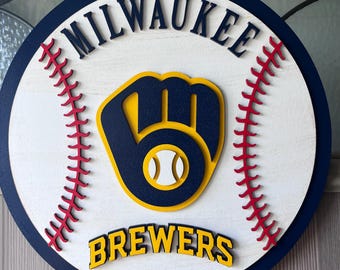 Könnte beinhalten: Rundes Holzschild mit dem Logo der Milwaukee Brewers. Das Schild hat einen weißen Hintergrund mit einem marineblauen Rand und roten Nähten. Das Brewers-Logo ist ein marineblauer und gelber Baseballhandschuh mit einem Baseball in der Mitte. Die Wörter "MILWAUKEE" und "BREWERS" sind zu sehen.