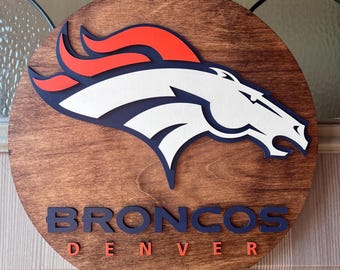 May include: A round wooden sign featuring the Denver Broncos logo. The logo is a white horse head with a blue outline and orange mane. The words "BRONCOS DENVER" are in blue and orange lettering.