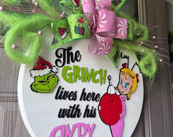 Puede incluir: Un letrero redondo de madera blanca con los personajes de The Grinch y Cindy Lou Who. El letrero tiene detalles en verde y rosa, incluyendo un lazo con un diseño de bastón de caramelo y el texto "The Grinch lives here with his Cindy Lou."