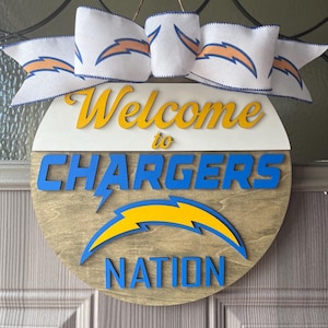 Könnte beinhalten: Ein rundes Holzschild mit den Worten "Welcome to Chargers Nation" in blauen und gelben Buchstaben. Das Schild hat einen weißen Hintergrund mit einem blauen und gelben Blitzlogo. Ein weißes Band mit blauen Blitzen ist um das Schild gebunden.