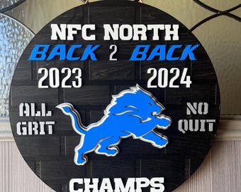 Könnte beinhalten: Ein rundes Schild aus schwarzem und weißem Holz mit einem blauen Löwenlogo. Das Schild zeigt "NFC North Back 2 Back 2023 2024 All Grit No Quit Champs".