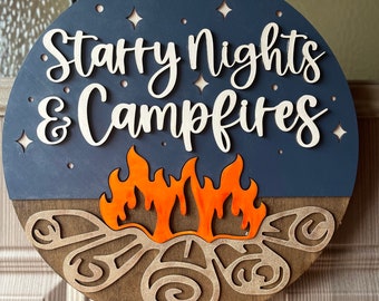 Könnte beinhalten: Runder Holzschild mit dem Schriftzug "Starry Nights & Campfires" in weißer Schrift. Das Schild zeigt einen dunkelblauen Himmel mit Sternaussparungen, eine Lagerfeuergrafik und einen braunen Sockel. Es hängt an einer natürlichen Schnur.