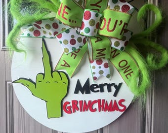 May include: A white round door hanger with a green Grinch hand giving the middle finger. The text "Merry Grinchmas" is written in black and red letters. A green, white, and red bow with polka dots and glitter is attached to the top.