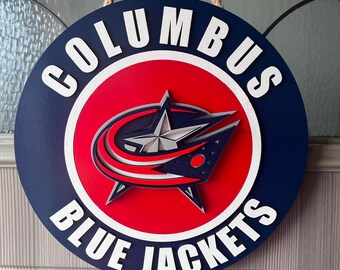 Puede incluir: Letrero redondo de madera con el logotipo de los Columbus Blue Jackets. El letrero tiene un fondo azul marino con las palabras "COLUMBUS" y "BLUE JACKETS" en blanco. El logotipo es un círculo rojo con una estrella y un diseño azul marino y plateado.