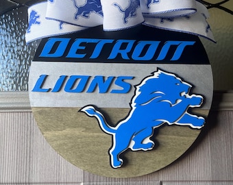 May include: A round wooden door hanger with a blue and white ribbon bow. The door hanger features a blue lion logo with the words "DETROIT LIONS" in black and white lettering.