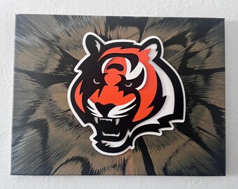 Könnte beinhalten: Ein Leinwandkunstwerk mit einem Tigerkopf-Logo in Orange, Schwarz und Weiß. Das Gesicht des Tigers steht im Mittelpunkt, vor einem Hintergrund mit einem strukturierten, federartigen Muster in Schwarz- und Brauntönen.