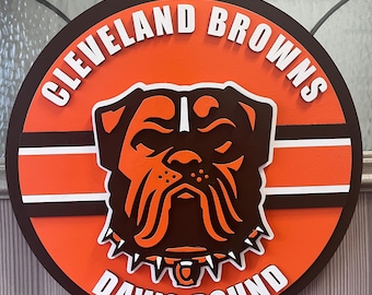 Könnte beinhalten: Rundes Holzschild mit dem Logo der Cleveland Browns. Das Schild hat einen orangefarbenen Hintergrund mit dem Teamnamen und "DAWG POUND" in weißer Schrift. Eine braune Bulldoggen-Grafik befindet sich in der Mitte, mit braunen und weißen Streifen.