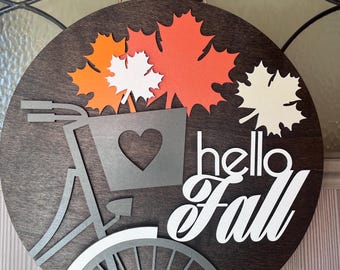 Könnte beinhalten: Ein rundes Holzschild mit einer grauen Fahrradsilhouette, einem Korb mit einem Herzen und Herbstblättern in Orange, Weiß und Rot. Das Schild trägt die Aufschrift "hello fall" in weißen Buchstaben.