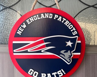 Könnte beinhalten: Rundes Holzschild mit dem Logo der New England Patriots. Das Schild hat einen roten Rand, eine marineblaue Mitte und weiße Schrift, die "NEW ENGLAND PATRIOTS" und "GO PATS!" lautet. Ein Seil ist zum Aufhängen befestigt.