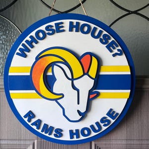 Puede incluir: Un letrero redondo de madera con el texto azul "WHOSE HOUSE? RAMS HOUSE". Tiene un fondo blanco con un logotipo de cabeza de carnero azul, amarillo y naranja. Colgando de una cuerda marrón.