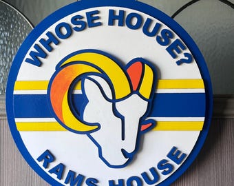 Könnte beinhalten: Rundes Holzschild mit dem blauen Text "WHOSE HOUSE? RAMS HOUSE". Es hat einen weißen Hintergrund mit einem blauen, gelben und orangefarbenen Widderkopf-Logo. Aufgehängt an einer braunen Schnur.