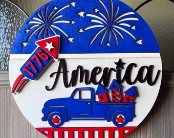 Könnte beinhalten: Ein runder Türhänger aus Holz mit patriotischem Thema. Der obere Teil ist blau mit Sternaussparungen und Feuerwerksdesigns. Das Wort "America" steht in schwarzer Schrift. Ein blauer Truck transportiert Feuerwerk.
